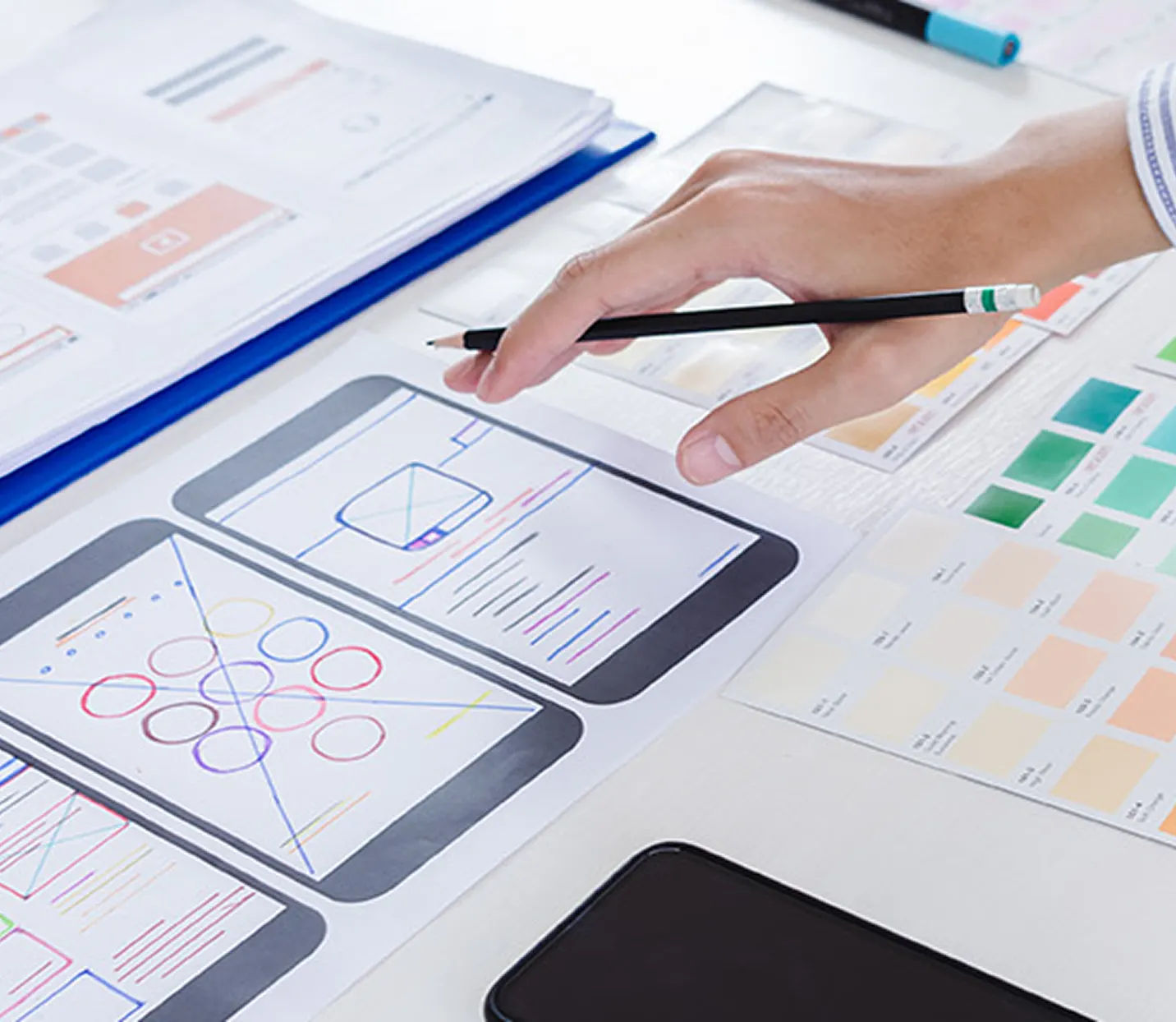 Hand holding pencil over a desk with printed mobile app wireframe sketches and color swatches.